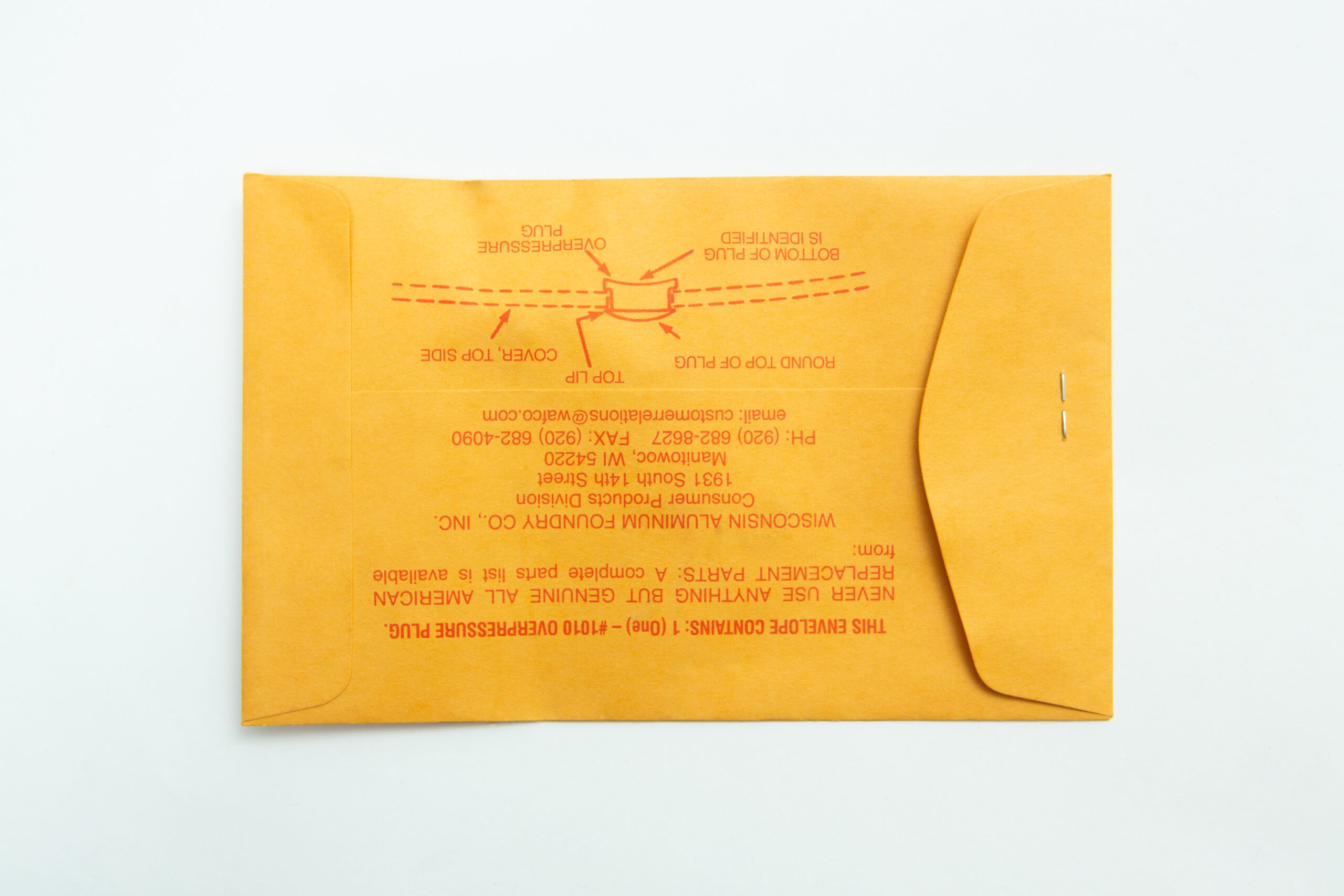 A yellow manila envelope with red instructions and a metal clasp lies on a white surface next to an orange All American rubber overpressure plug for sterilizer, Part 1010.