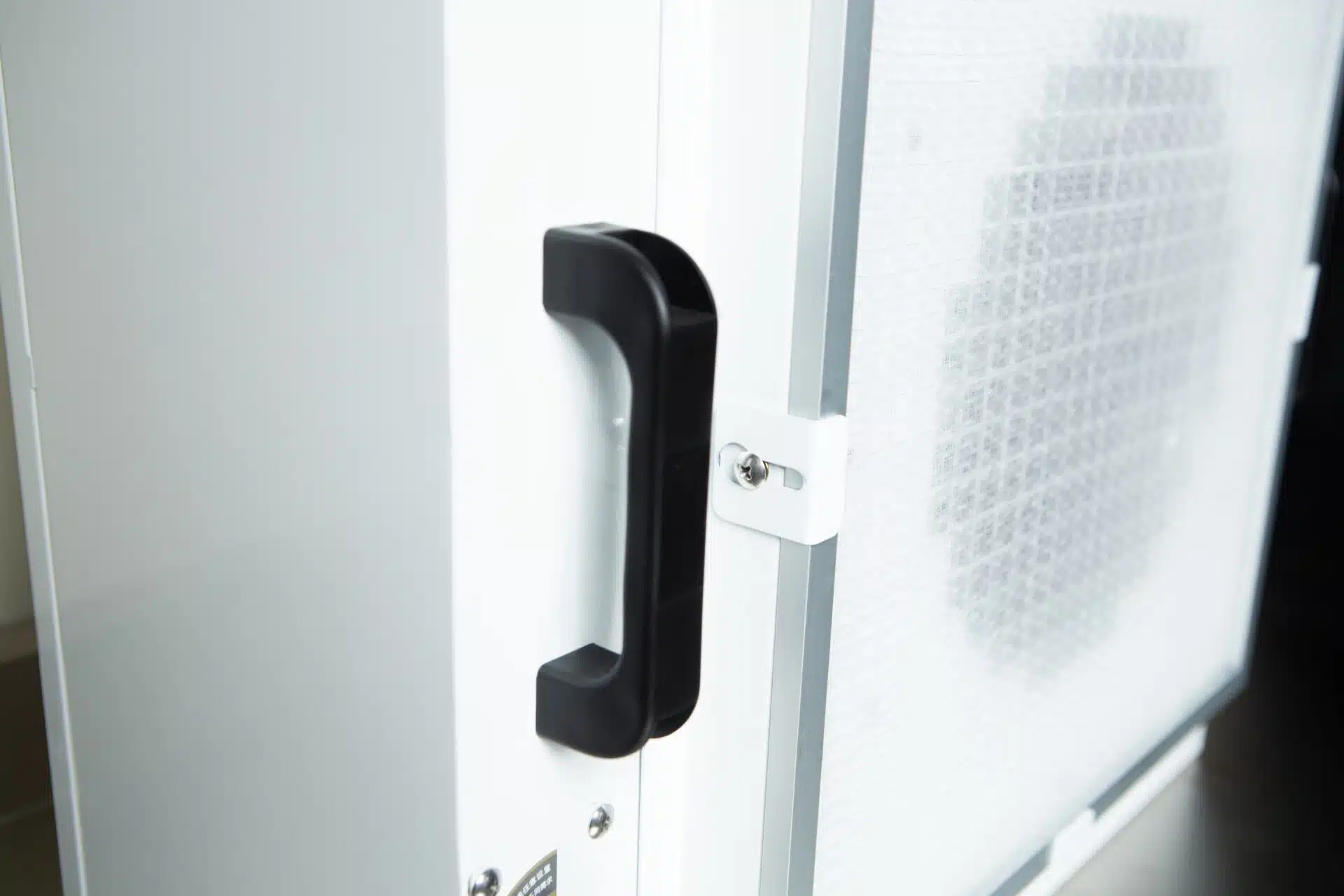 Close-up of a black handle on a white door next to the mesh panel of the Laminar Flow Hood HEPA Airfilter H14 w/prefilter 60x60x35cm.