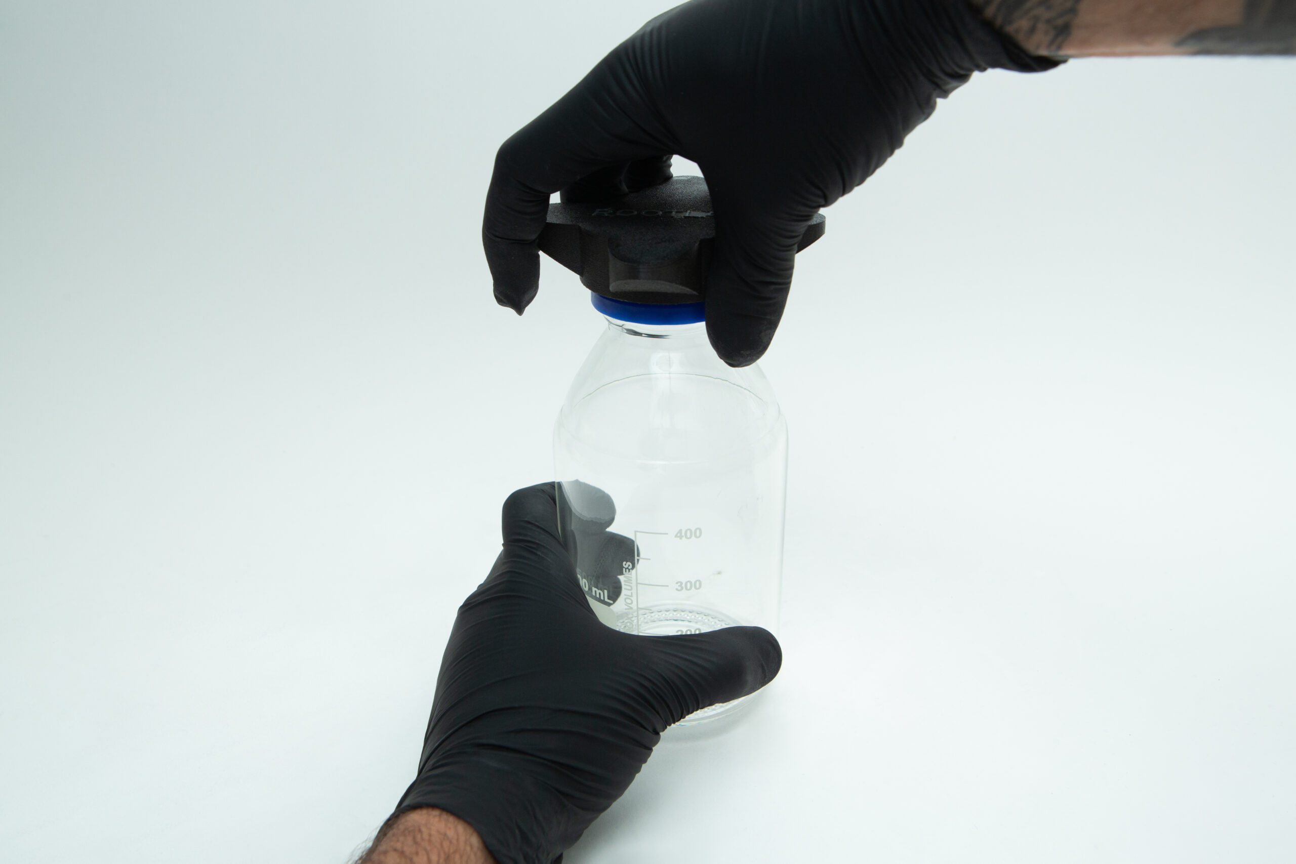 Gloved hands holding a clear glass jar with a black lid against a white background.