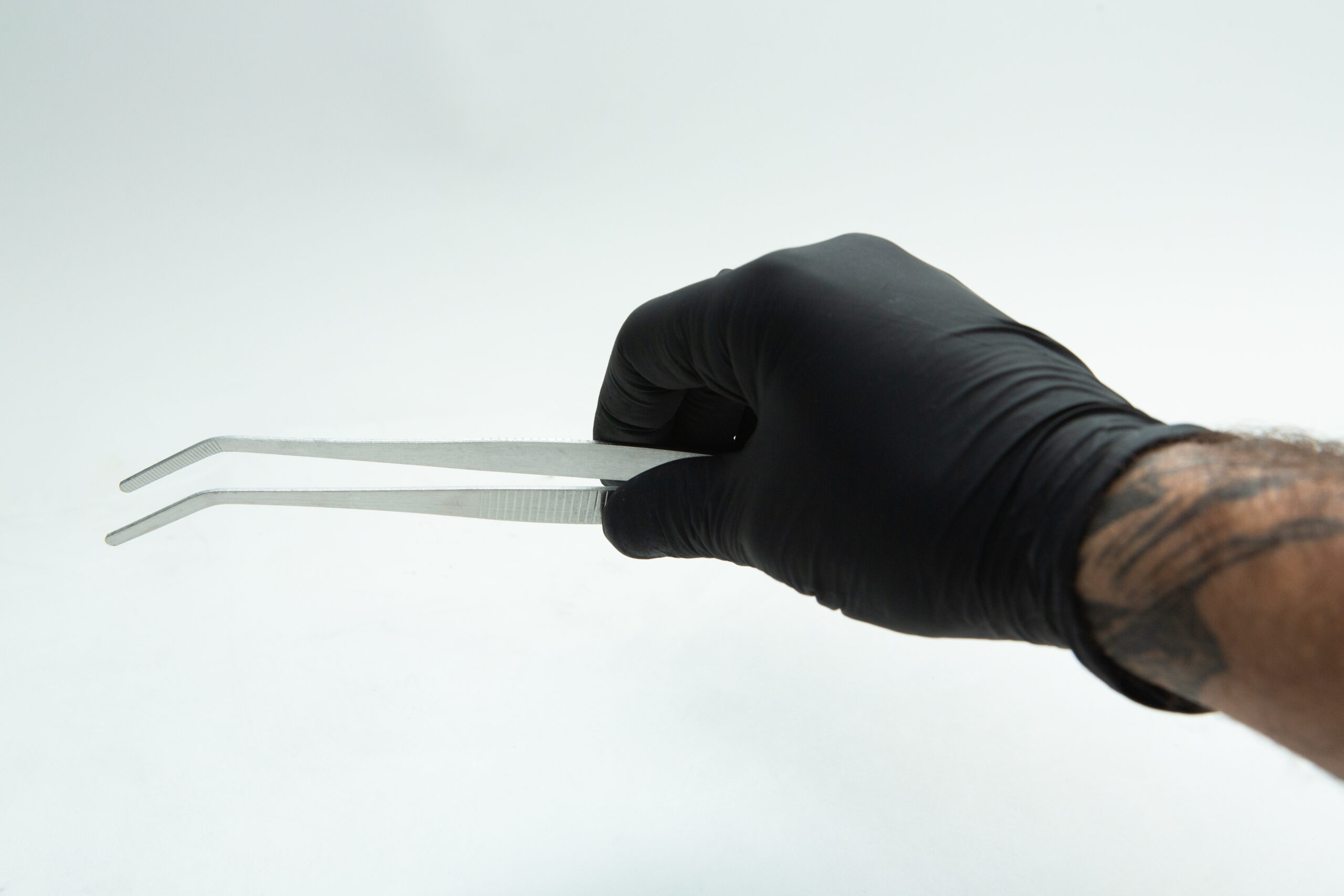 A hand wearing a black glove holds two metal tweezers against a plain white background.
