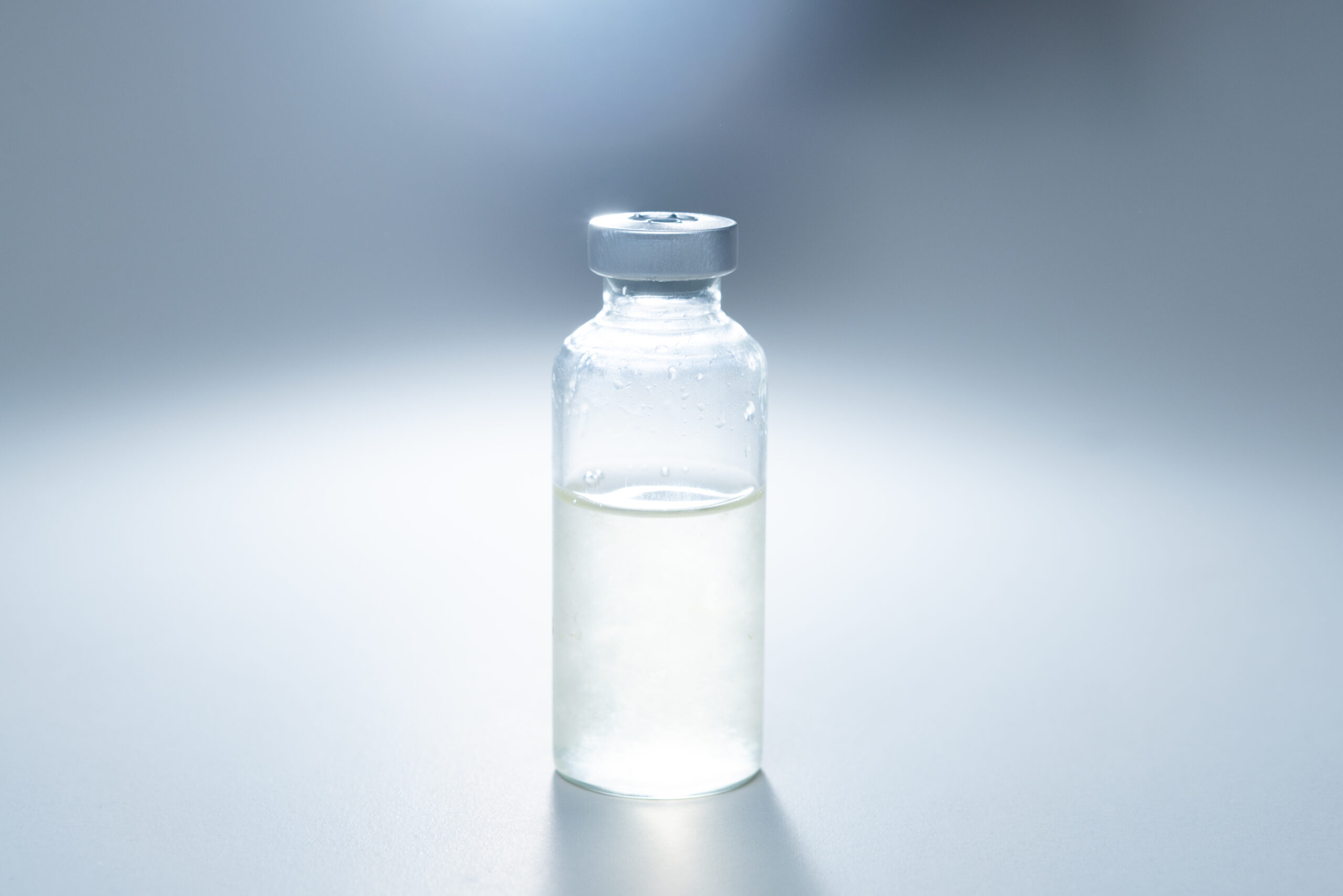 A small glass vial filled halfway with clear liquid, standing on a plain white surface under bright light.