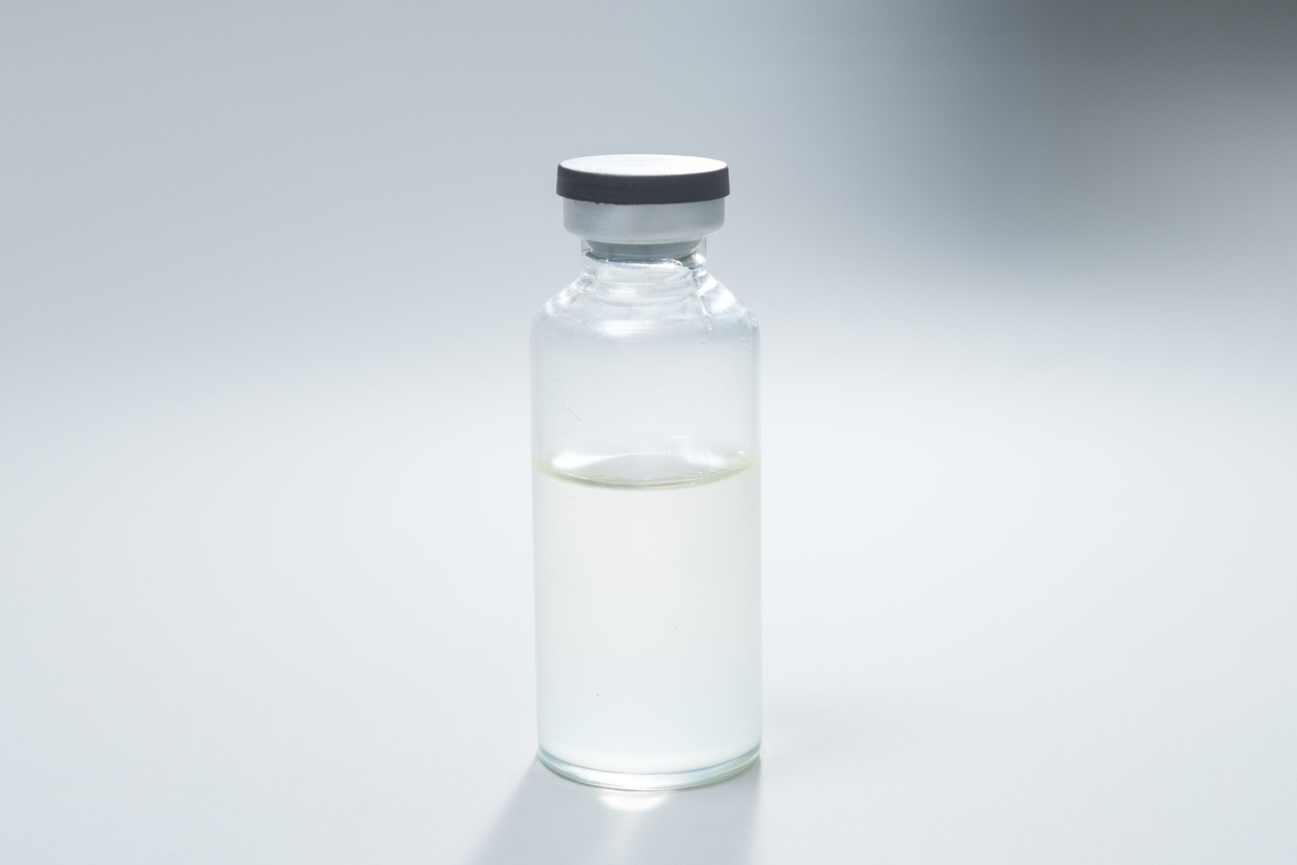 A small glass vial with a gray cap, partially filled with a clear liquid, sits on a white surface.