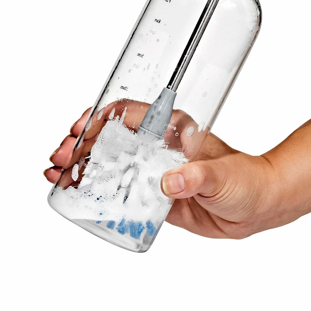 Hand cleaning a clear bottle with a soapy brush inside, visible bubbles and water droplets.