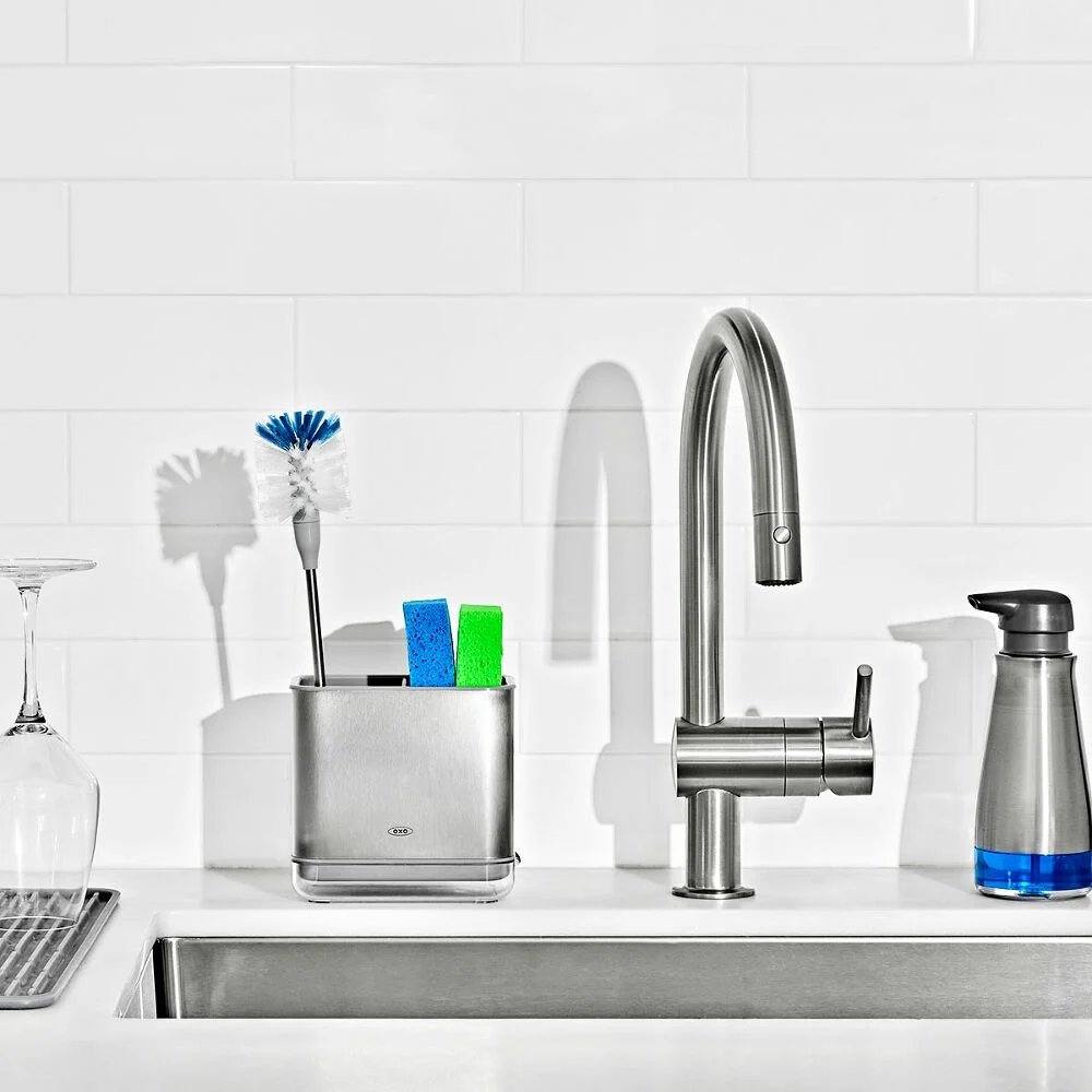 A kitchen sink with a faucet, soap dispenser, sponge holder, glass, sponges, and a brush on a white background.