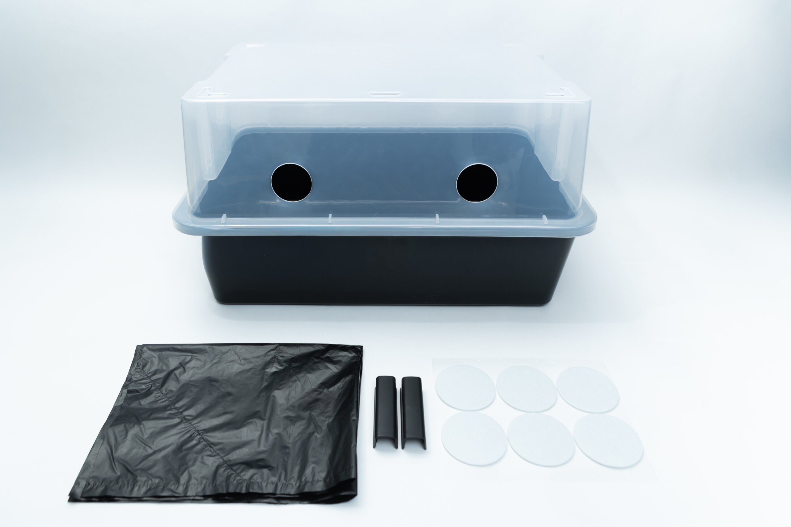 A plastic storage bin with two circular side holes, accompanied by a black liner, two clips, and six adhesive filters.